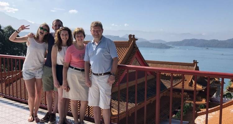 Smiling travellers stand on a terrace overlooking ornate temple rooftops and a wide blue lake with mountains.