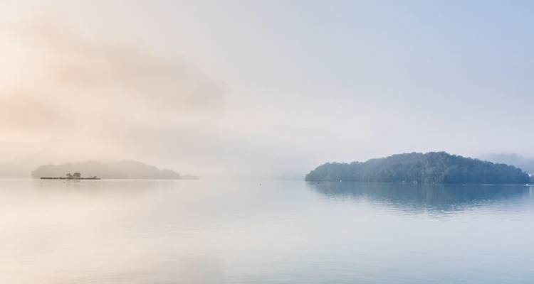 Misty sunrise view of small forested islands floating on a calm lake with soft pastel sky.