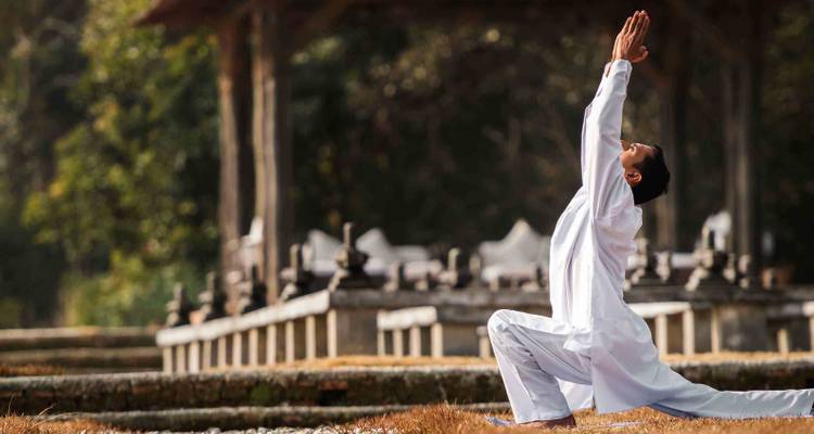 Un hombre vestido de blanco practica yoga en un jardín al aire libre cerca de estructuras de piedra antigua.