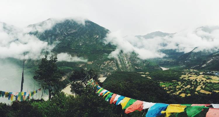 Banderas de oración coloridas extendiéndose a través de un valle brumoso con montañas envueltas en nubes y tierras de cultivo.
