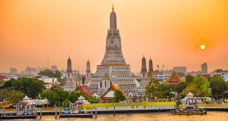 Der Prang des Wat Arun erhebt sich über den Chao-Phraya-Fluss unter einem orangefarbenen Abendhimmel.