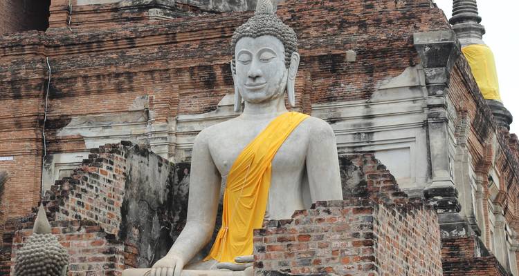 Weathered stone Buddha clad in yellow sash sits against ancient brick pagoda.