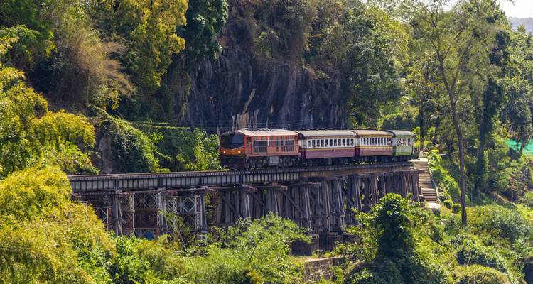 Train crosses historic wooden trestle bridge through lush jungle terrain.