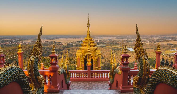 Golden mountaintop temple with naga balustrades overlooking rural plains at dawn.