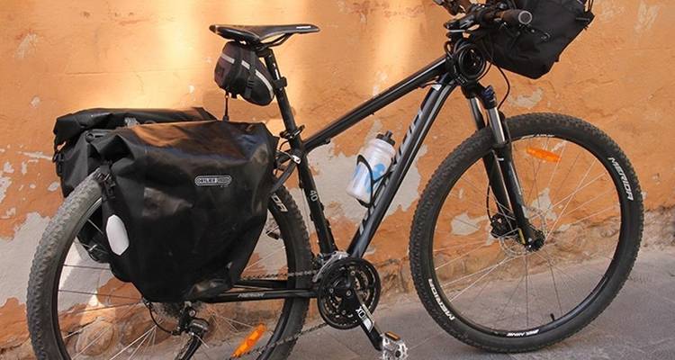 Close-up van een volgeladen toerfiets met tassen tegen een getextureerde okergele muur.