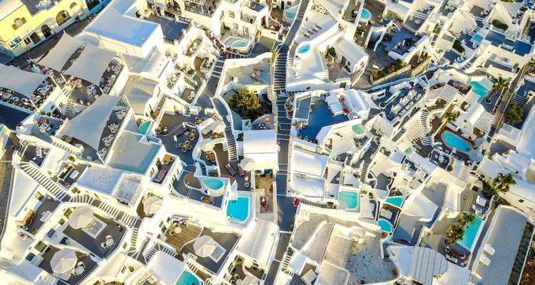 Drone view of tightly packed whitewashed dwellings with turquoise pools on Santorini cliff