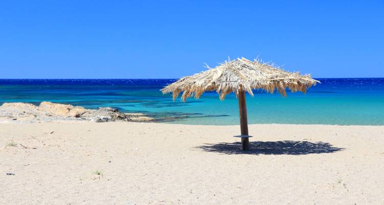 Single straw beach umbrella casting shadow on empty sandy shore beside turquoise sea