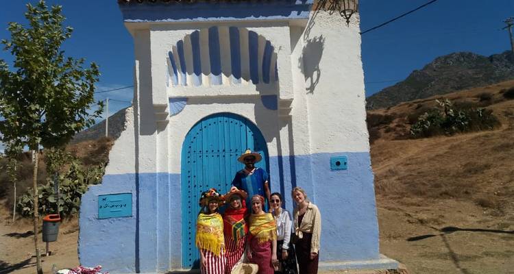 Des voyageurs en châles colorés posant devant une porte cintrée blanche et bleue dans un cadre rural en montagne.