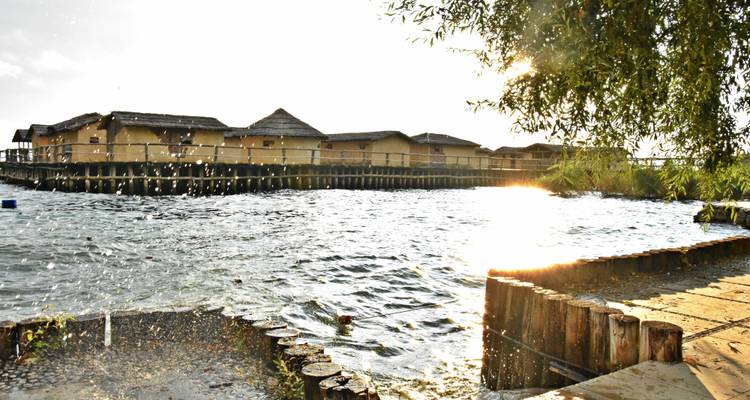 Vue sur le lac avec des maisons traditionnelles sur pilotis et un soleil éclatant se reflétant sur l'eau.