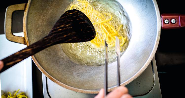 Primer plano de fideos fritos en aceite en un wok, espátula de madera y pinzas en acción.
