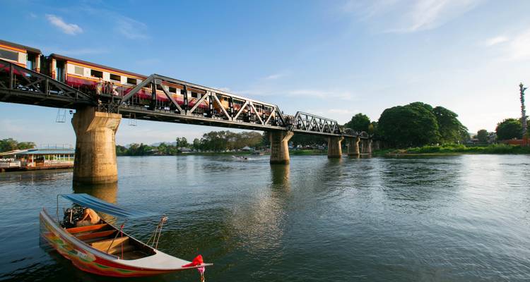 Puente histórico sobre el río Kwai con un tren cruzando sobre aguas tranquilas; bote de cola larga en primer plano.
