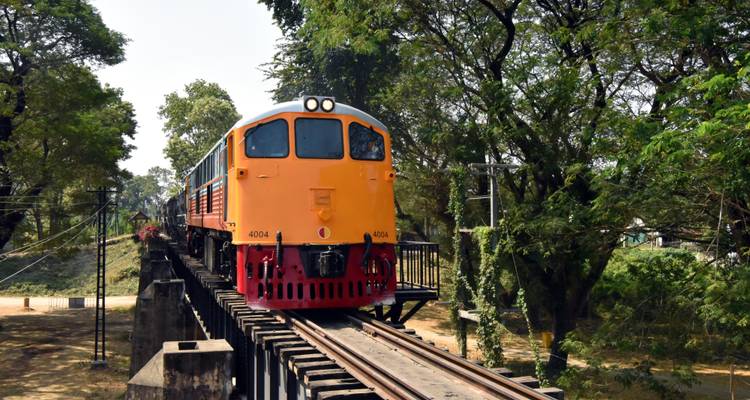 Locomotora naranja cruzando un puente de madera a través del Ferrocarril de la Muerte en medio de un exuberante bosque.