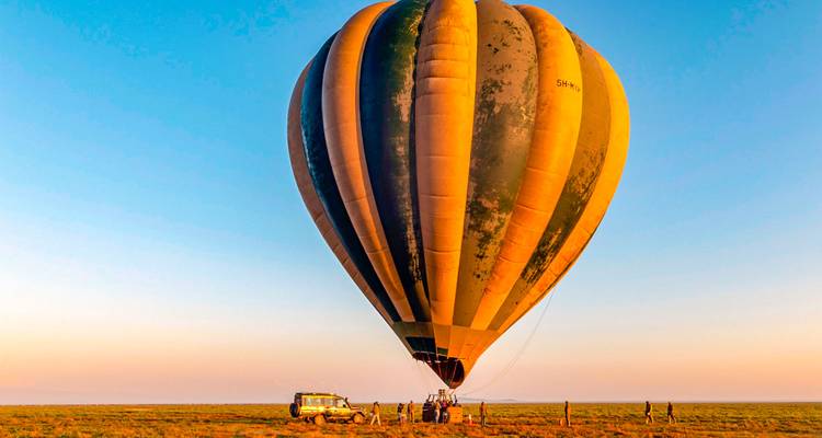 Giant striped hot air balloon tethered on open plain at sunrise with crew and safari vehicle nearby
