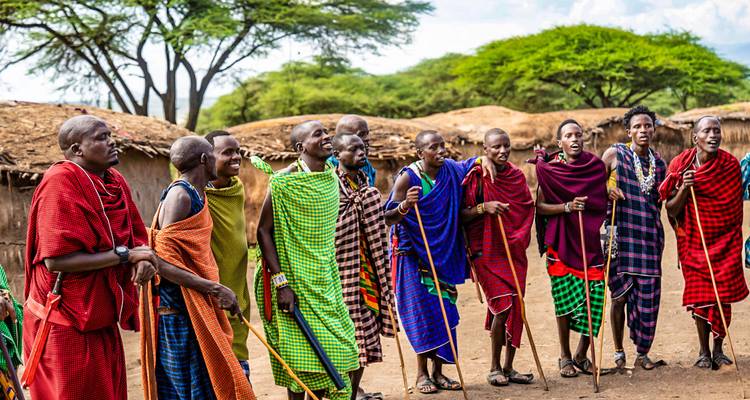 Maasai men and women sing together outside earthen huts amid acacia trees