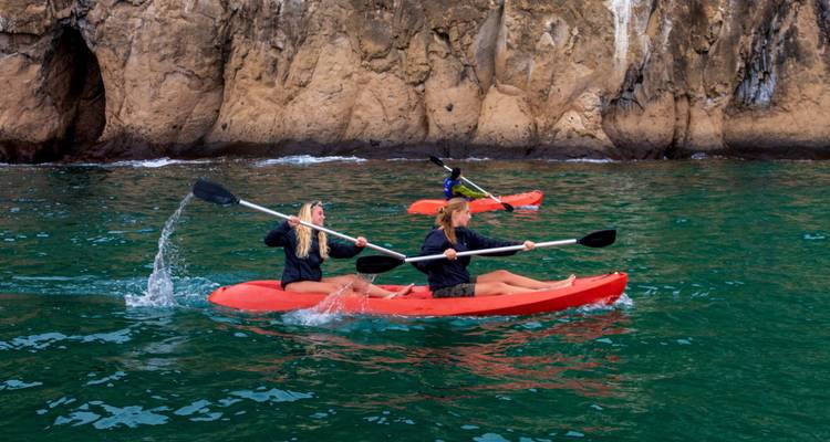 Two people kayaking in clear waters with rocky cliffs in the background.