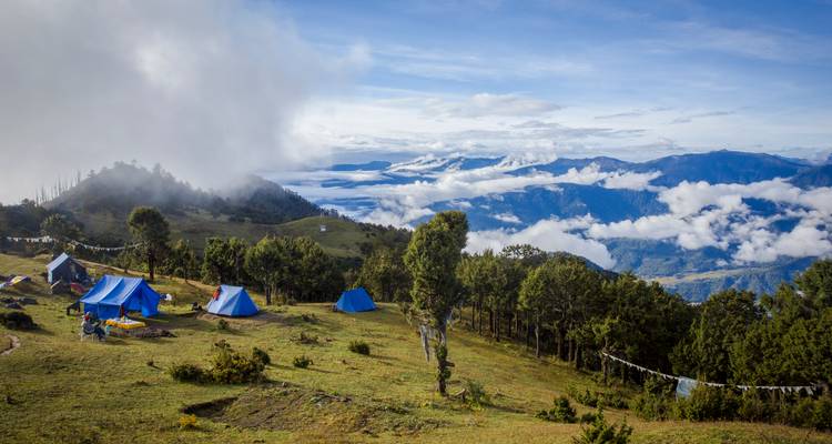 Un campement sur une crête herbeuse parsemée de tentes bleues surplombe des nuages qui roulent, des collines lointaines et des drapeaux de prière.