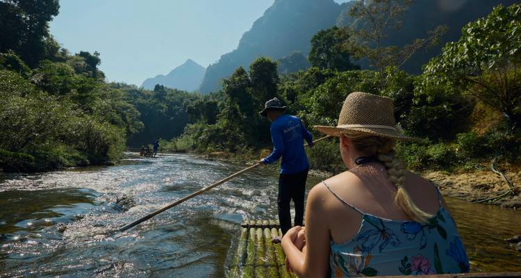 Tourist sits on bamboo raft while guide poles it through a sunlit jungle river valley.