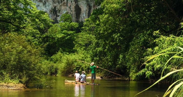Small group glides on a bamboo raft through lush green jungle with limestone cliffs in the background.