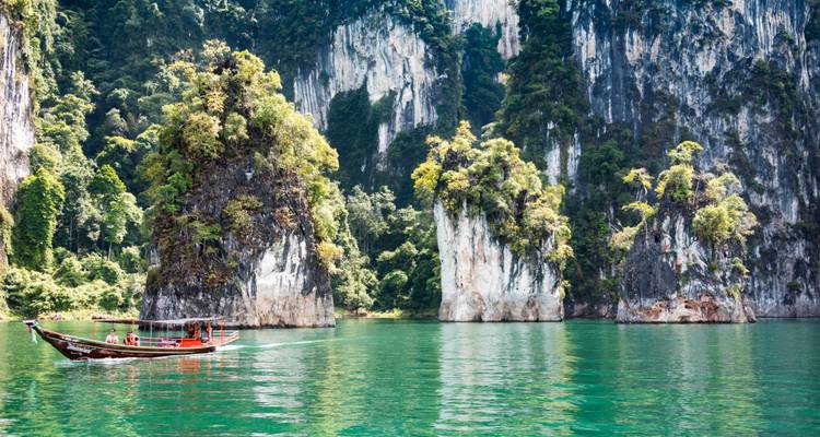Long-tail boat cruises on emerald lake beside towering jungle-clad cliffs of Khao Sok National Park.
