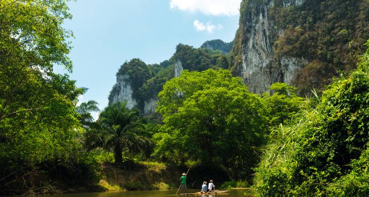 Bamboo raft drifts down jungle river beneath steep limestone cliffs and lush vegetation.