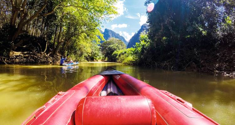 View from a red inflatable kayak gliding down a jungle river toward distant limestone peaks.