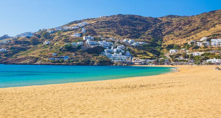 Sandy beach with clear blue water and houses on a hillside.