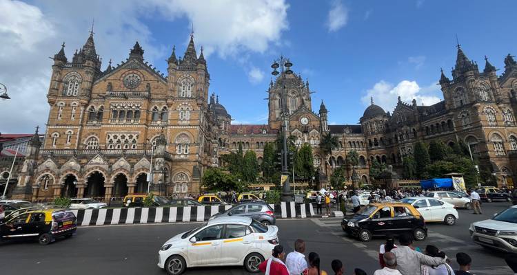 A bustling city street in front of a historic railway station with traffic and pedestrians.