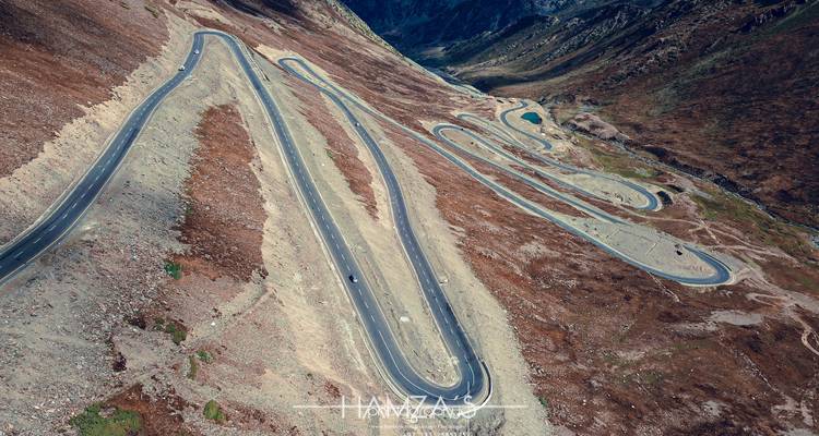 Drone-opname van een kronkelende bergweg met talrijke haarpeldboches die door kale rotsachtige hellingen snijden.