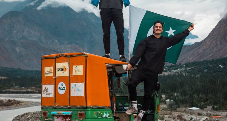 Reizigers staan op een kleurrijke tuk-tuk en zwaaien met een Pakistaanse vlag met dramatische bergen op de achtergrond.