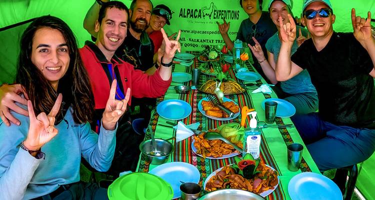 Gruppe von Trekking-Enthusiasten in einem grünen Speisezelt, die auf Tellern servierte Mahlzeiten genießen und freudig gestikulieren