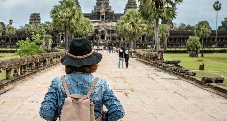 Young traveler with a backpack and black hat walking along the stone causeway toward Angkor Wat temple surrounded by palms and lawns.