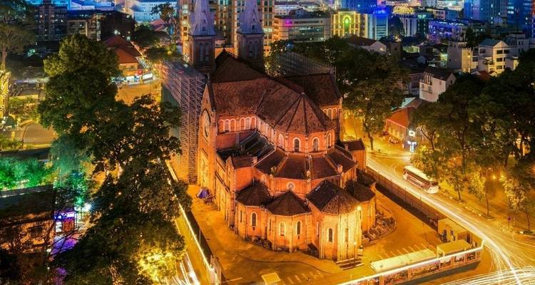 Night aerial view of the illuminated red-brick Notre-Dame Cathedral in Ho Chi Minh City with traffic light trails and city skyline.