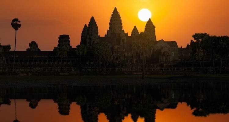 Silhouette of Angkor Wat reflected in still water under a glowing orange sunrise sky with the sun above the towers.