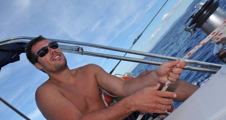 Eine Person mit Sonnenbrille an Bord eines Segelboots, die sich auf das Handhaben von Seilen konzentriert.