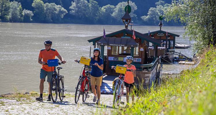 Family pushes their bicycles up a cobbled ramp toward a small Danube ferry on a sunny riverside trail.