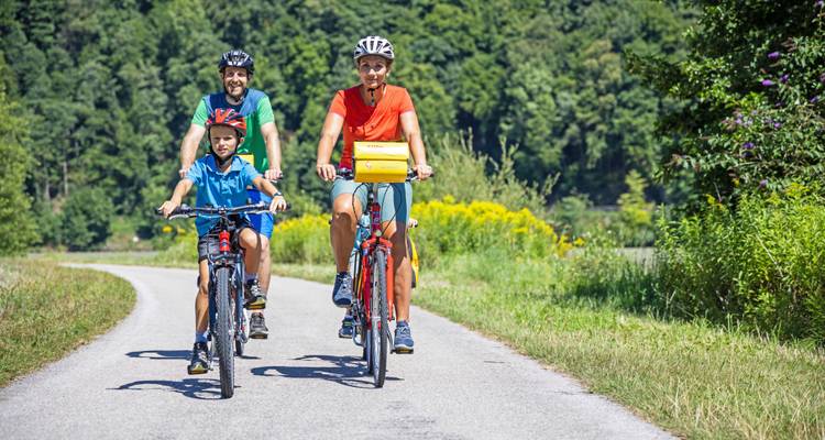 Parents and child ride bicycles along a winding rural path bordered by lush greenery.