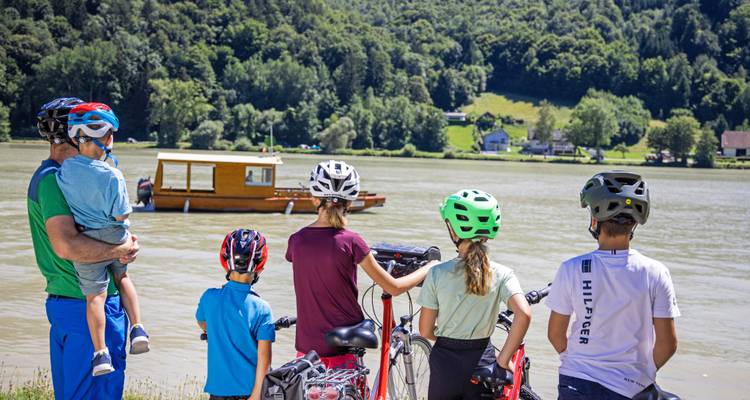 Children and adults watch a wooden boat on the Danube while standing beside their bikes.