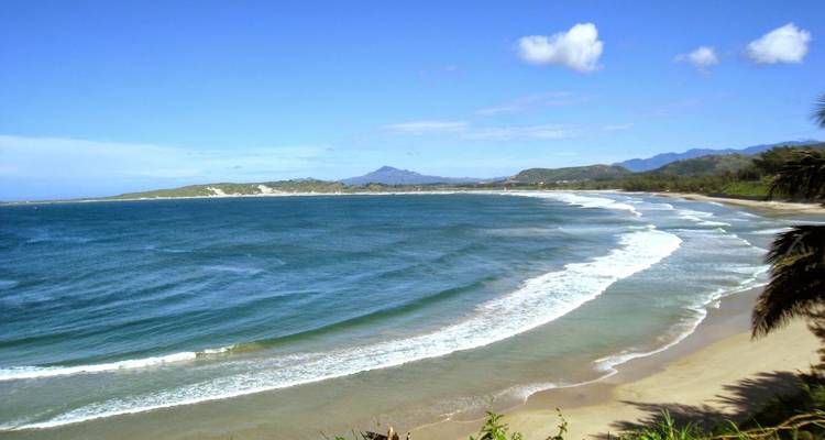 Prachtige kustlijn met golven die op een zandstrand beuken, blauwe lucht en bergen op de achtergrond.