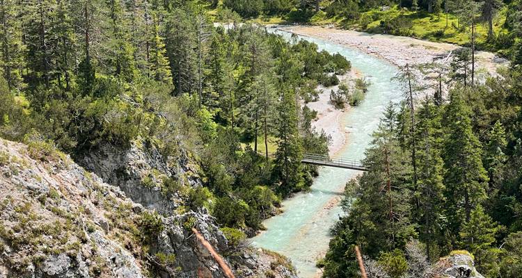 Rivière de montagne turquoise serpentant à travers une gorge forestière escarpée avec un étroit pont à pied en bois la traversant.