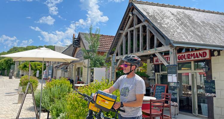 Un cycliste en tournée s'arrête devant un café rustique et une boutique gastronomique sur une rue ensoleillée de la vallée de la Loire.