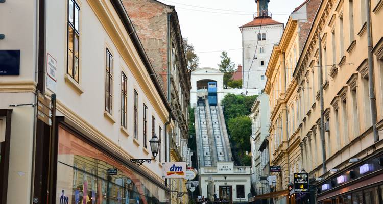 Rue animée du centre-ville de Zagreb menant au funiculaire historique et à la tour avec les fils de tramway surplombant la scène.