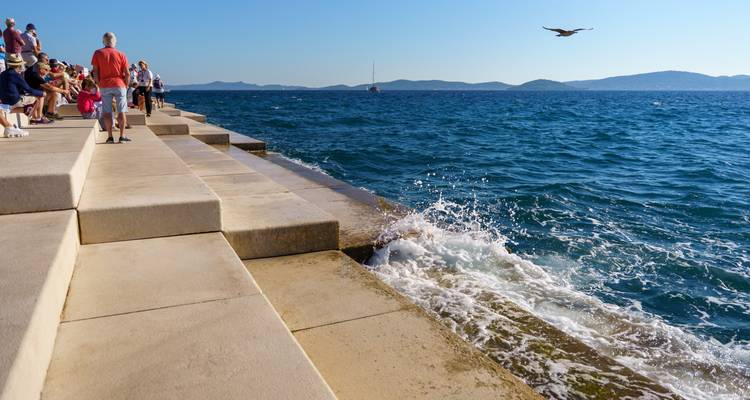 Les visiteurs s'assoient sur les marches en béton de l'Orgue de la Mer de Zadar et regardent les vagues s'écraser et projeter des embruns sous un ciel dégagé.