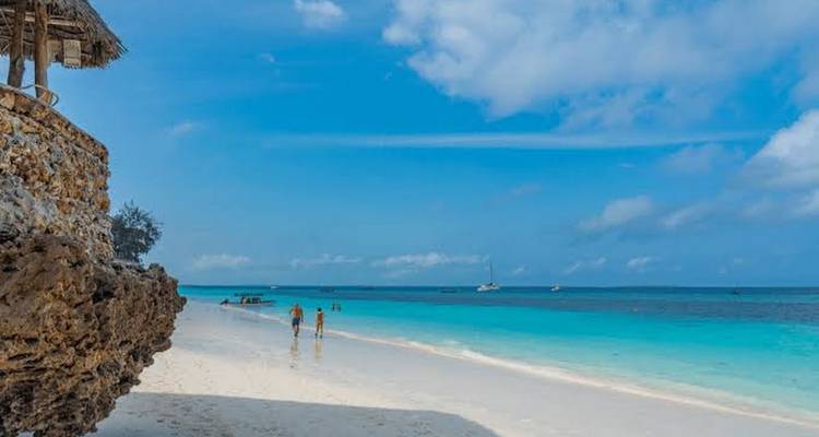 Beach scene with people walking near crystal-clear water and a sailboat in the distance.