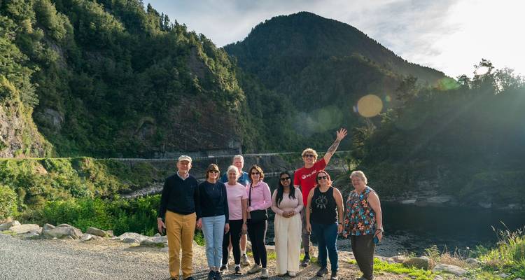 Grupo de turismo sonriente posando junto a un río con montañas boscosas y destello de sol por la tarde.