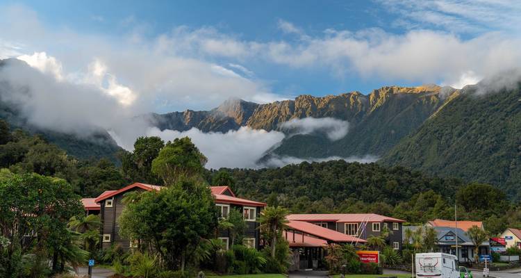 Pequeño pueblo alpino acurrucado bajo montañas dentadas y brumosas con nubes bajas por la mañana.