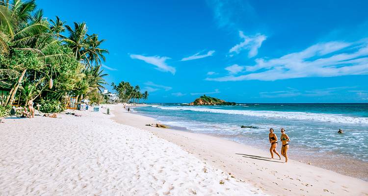 Eine tropische Strandszene mit Touristen, die bei strahlendem Himmel am Ufer entlanggehen.