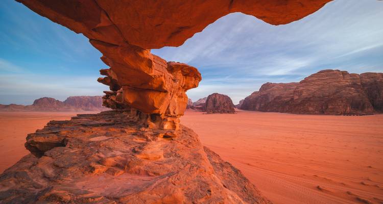 Felsenbrückenformation erhebt sich über orangefarbene Sandebenen unter sanftem Morgenhimmel im Wadi Rum.