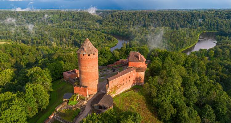 Aerial view of red-brick Turaida Castle rising above dense green forest with winding river below.