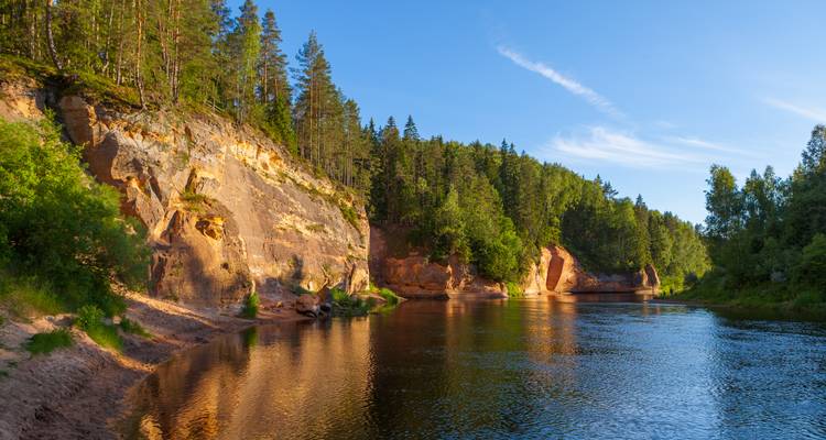 Serene river flowing past golden sandstone cliffs and pine forests under a clear blue evening sky.