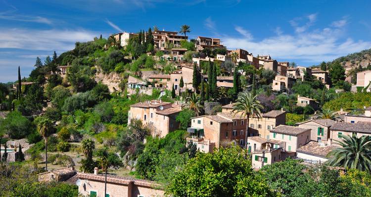 Pintoresco pueblito en la ladera con casas de piedra, palmeras y cipreses bajo un cielo azul brillante.